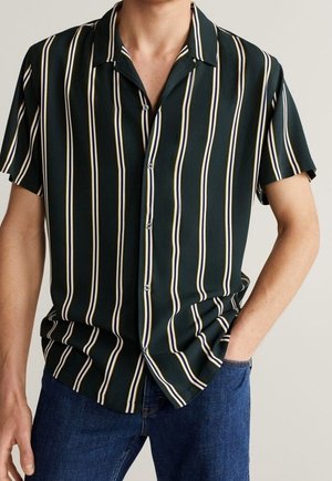 Uomo con camicia verde scuro a maniche corte con righe verticali bianche e beige, abbinata a jeans di denim blu, mano in tasca.