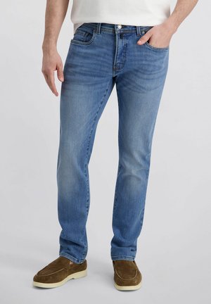 Mężczyzna w jasno-niebieskich dopasowanych dżinsach, białej koszuli i brązowych zamszowych wsuwanych butach, stojący z jedną ręką w kieszeni.