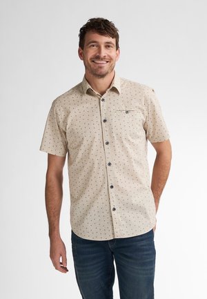 Beige Kurzarmhemd mit kleinen gemusterten Punkten, Button-Down-Kragen und Brusttasche; kombiniert mit blauen Jeans.