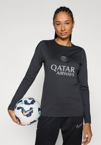 Fekete hosszú ujjú sportfelső "Qatar Airways" logóval, kerek nyakkivágással és nedvességelvezető anyagból, fekete nadrággal viselve, és focilabdát tartva.