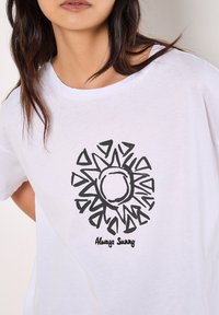 Weißes Baumwoll-T-Shirt mit einem schwarzen Sonnenmotiv mit gezackten Strahlen und dem Text "Always Sunny" unterhalb des Designs. Kurzarm, lässige Passform.
