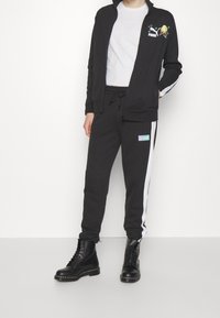 Personne portant un survêtement Puma noir avec des bandes blanches sur les côtés, arborant un motif SpongeBob sur la veste, assorti à des bottes noires à lacets.