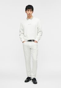 Λευκό φούτερ με κόκκινο λογότυπο "Michael Kors", μπεζ πουκάμισο με γιακά από μέσα, συνδυασμένο με λευκό παντελόνι και μαύρα loafers.