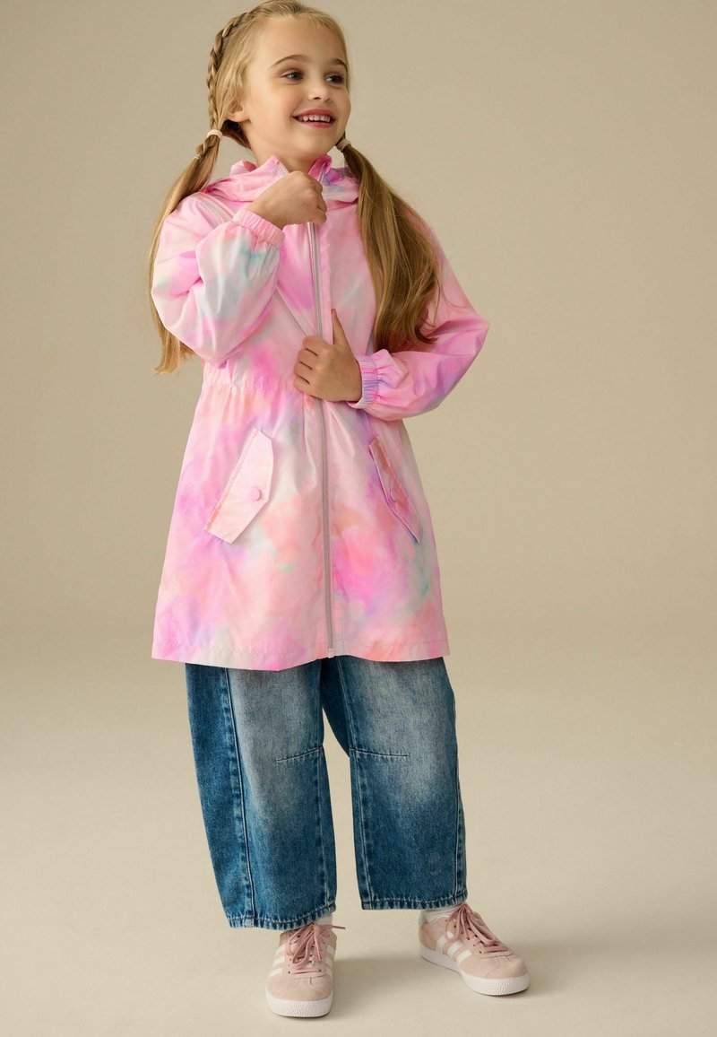 Junges Mädchen lächelt, trägt eine rosa Batik-Kapuzenjacke, weite blaue Jeans und hellrosa Turnschuhe, steht vor einem einfarbigen Hintergrund.