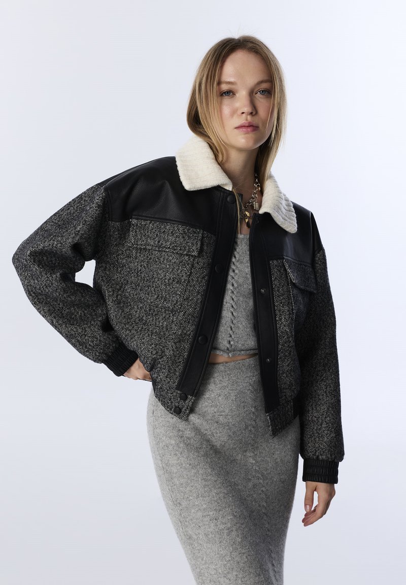 Vrouw in een grijze gebreide rok en crop top met een zwart-grijze wollen jas met leren accenten en een witte kraag, poseert voor een witte achtergrond.