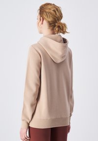 Sudadera beige con bolsillo canguro y capucha con cordones. Textura suave, puños y dobladillo acanalados. Lleva puestos unos pantalones burdeos.