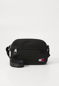 Zwarte rechthoekige crossbodytas gemaakt van duurzaam stof, met een ritssluiting aan de voorkant, verstelbare riem en een logopatch in rood, wit en blauw.