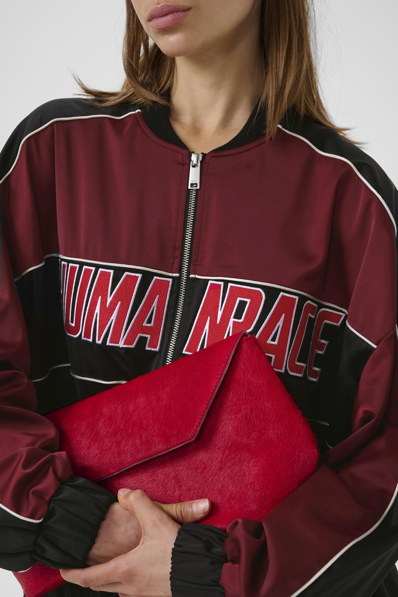 Woman wearing a maroon and black Puma Race jacket holding a large red envelope clutch bag against a white background.