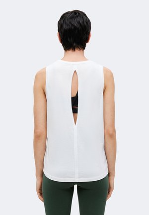 Personne aux cheveux courts et foncés porte un haut blanc sans manches avec une fente verticale dans le dos, révélant un soutien-gorge de sport noir en dessous et des leggings verts.