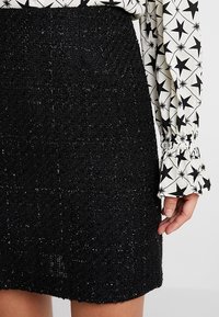 Svart texturerad kjol med subtila metalltrådar, med rak skärning och midjeresår, kombinerad med en mönstrad blus med långa ärmar.