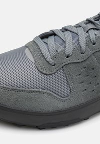 Sivi športni čevelj, ki vključuje kombinacijo mrežaste in semiš usnjene materiala, teksturiran dizajn, ravne vezalke ter gumijast podplat z ventilacijskimi luknjami.
