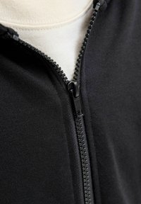 Sweat à capuche zippé noir en matière lisse. Présente une fermeture à glissière visible et une superposition d'un tissu couleur crème en dessous.