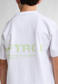 Bílá bavlněná tričko s krátkým rukávem, na zadní straně s jemným zeleným grafickým textem, který zní "FTRC Pedustries International D Beach."