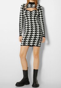 Svartvit klänning i hounds-tooth-mönster med långa ärmar, urklippt halsringning och figurnära design. Bärs med svarta ankelskor.