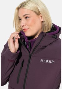 Chaqueta impermeable morada con un alto cuello, cremalleras negras y cordones ajustables. Presenta un logo en el pecho y un forro interior morado.