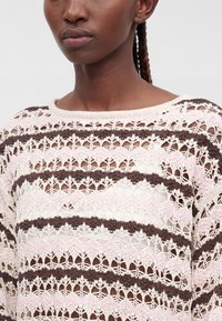 Femme aux cheveux tressés portant un pull en crochet à rayures horizontales blanc, rose clair et marron.
