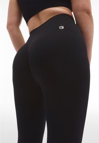 Leggings neri con vita alta, texture liscia e delicato accento del logo. La vestibilità si adatta al corpo, evidenziando le curve.