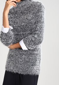 Maglione grigio e bianco fluffy con texture, colletto alto e maniche a tre quarti, indossato sopra una camicia bianca con colletto.