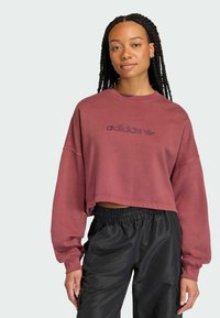 Felpa corta bordeaux con scollo rotondo e spalle scese, caratterizzata da un logo testurizzato in bordeaux più scuro. Abbinata a pantaloni neri.