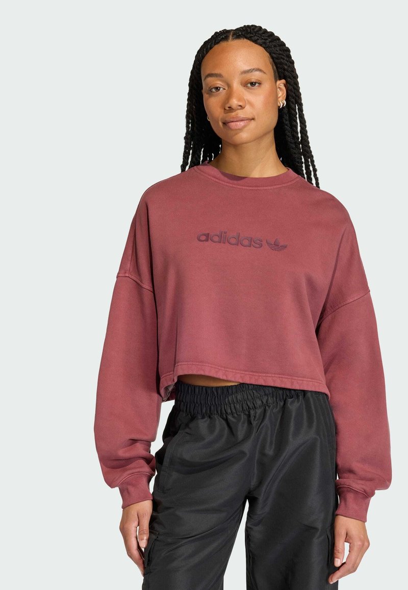 Felpa corta bordeaux con scollo rotondo e spalle scese, caratterizzata da un logo testurizzato in bordeaux più scuro. Abbinata a pantaloni neri.