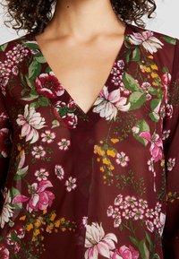 Kobieta w burgundowej bluzce z dekoltem w kształcie litery V, zdobionej różowymi, białymi, żółtymi i zielonymi kwiatowymi wzorami, z widocznymi kręconymi ciemnymi włosami.