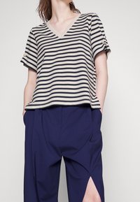 Camiseta de cuello en V a rayas en color beige y marino con mangas enrolladas, que cuenta con botones decorativos. Combinando con pantalones marinos con aberturas laterales.