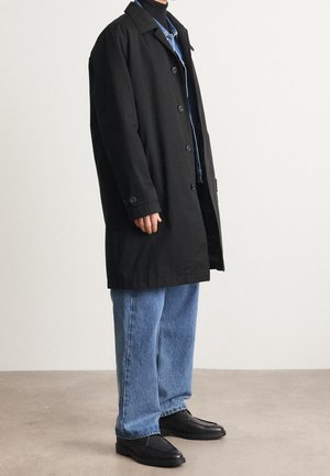 Personne portant un long manteau noir boutonné par-dessus un jean bleu et des chaussures noires, debout sur un sol uni devant un mur blanc.