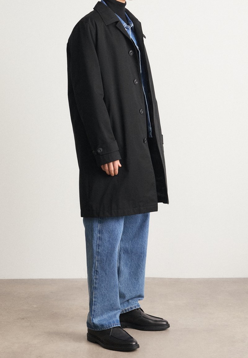 Personne portant un long manteau noir boutonné par-dessus un jean bleu et des chaussures noires, debout sur un sol uni devant un mur blanc.