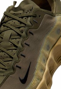 Oliivinvihreä urheilukenkä, jossa on teksturoitu kangaspäällinen, musta Nike-logo ja aaltoilevat koristeet. Kevyt pohja kuvioidulla pidolla.