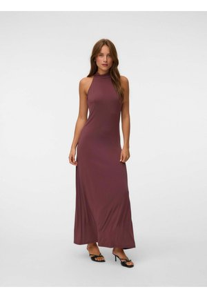 Woman stands wearing a sleeveless, high-neck mauve maxi dress with black open-toe heels on a plain light gray background.
