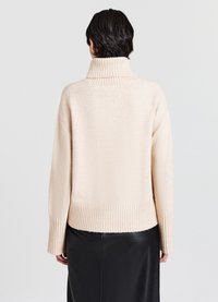 Beige Rollkragensweater aus weichem Material, mit geripptem Kragen und Saum, lockerer Passform und leicht abgesenkten Schultern. Eine schwarze Lederrock ist sichtbar.
