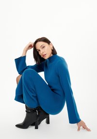 Langärmliges turkises Kleid mit hohem Kragen, weicher Textur und fließender Silhouette, kombiniert mit schwarzen Ankle Boots mit quadratischem Absatz.