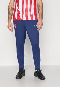 Blå træningsbukser i slim fit med et gult Nike-logo og Atlético de Madrid-logo, parret med sorte sportsko.