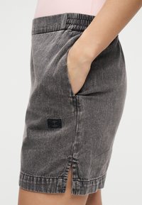 Χέρι στην τσέπη φούστας από ξεθωριασμένο μαύρο τζιν με πλαϊνή σχισμή και ελαστική μέση, φορεμένη με ανοιχτό ροζ τοπ.