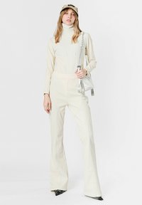 Mono ajustado color crema con cuello alto y piernas acampanadas, hecho de tela suave. Los accesorios incluyen un bolso y una gorra de color claro.