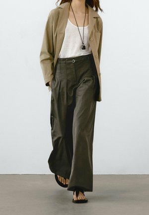 Personne portant un blazer beige, un haut blanc, un pantalon large foncé, des sandales noires et un long collier pendentif noir sur un fond clair uni.