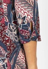 Ręka w wzorzystym rękawie z tkaniny w kolorze bordowym, granatowym i białym, z kwiatowymi i geometrycznymi wzorami, zapinana na guzik.