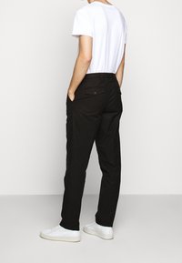 Muž v černých kalhotách s knoflíkovými zadními kapsami, bílém tričku a bílých teniskách, stojící na jednobarevném světlém pozadí.