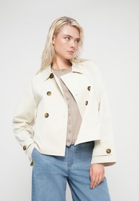 Veste courte couleur crème en matière lisse, avec de gros boutons marron, un col classique et deux poches latérales.