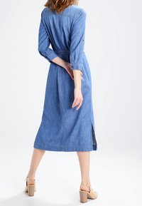 Nainen keskipitkässä farkkutakissa, jossa on kolme neljännesosaa hihat, ja beiget paksupohjaiset sandaalit, seisoo yksi käsi selän takana.