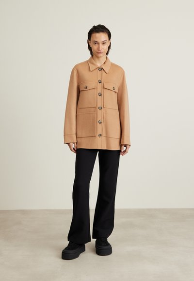 Hellbraune Wollmischung Jacke mit großen Fronttaschen, Knopfverschlüssen und klassischem Kragen. Kombiniert mit schwarzen Hosen und Plateauschuhen.