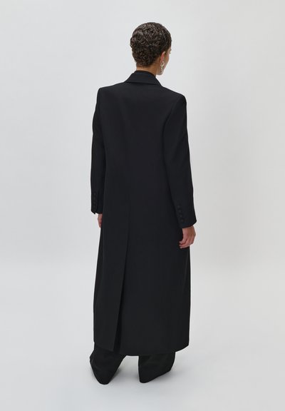 Persona con capelli corti e ricci indossa un lungo cappotto nero con spacco posteriore, pantaloni neri e orecchini d'argento, in piedi contro uno sfondo chiaro e uniforme.