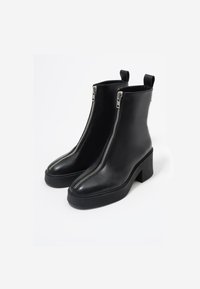 Botas negras de tobillo hechas de cuero suave con un alto tacón cuadrado y detalle de cremallera plateada expuesta, con una punta redondeada y lengüetas para tirar.