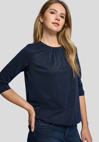 Dunkelblaues Langarmoberteil aus strukturiertem Stoff mit rundem Ausschnitt und gerafftem Detail am Kragen. Kombiniert mit dunklen Jeans.