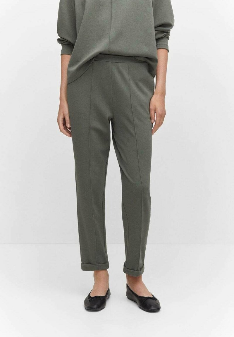 Personne portant un pantalon slim vert olive avec des revers roulés, un haut assorti et des ballerines noires, debout devant un fond uni.