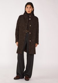 Cappotto marrone con texture, colletto alto, bottoni frontali e spacchi laterali, abbinato a pantaloni a righe scure e scarpe stringate marroni.