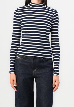 Frau trägt ein dunkelblau-weiß gestreiftes, langärmliges Rollkragenoberteil und dunkelblaue Denim-Jeans, steht vor weißem Hintergrund.