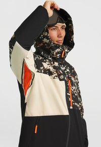 Skijack met een zwart en beige patroon, voorzien van oranje accenten, een capuchon en ritszakken. Textuur, duurzaam materiaal.