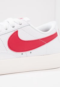 Vita Nike-sneakers med en texturerad läderöverdel, framträdande röd logga och vit gummisula. Klassisk lågskor design med snörning.
