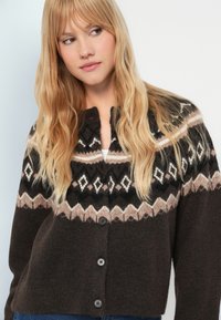Cardigan brown cu un model geometric în crem și bej, având front butonat, textură moale și mâneci lungi.